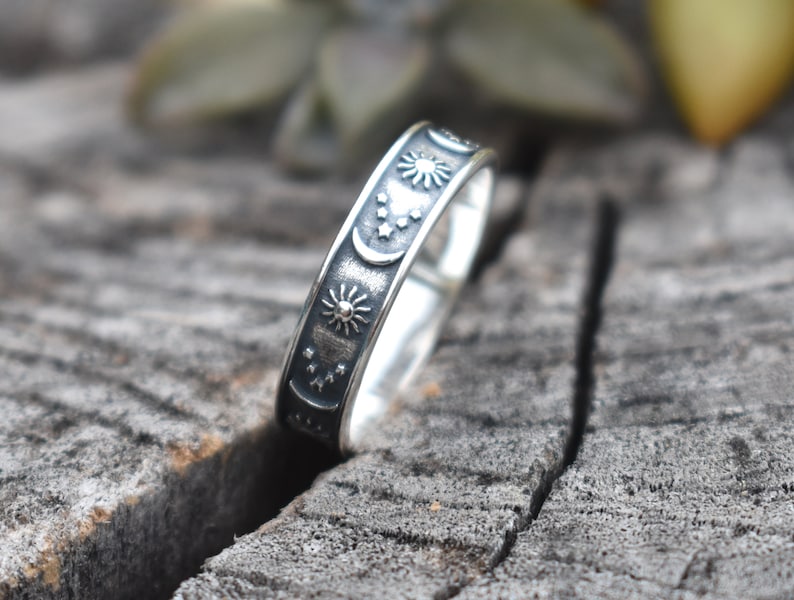 May include: A silver ring with a celestial design featuring moons, stars, and suns. The ring is on a wooden surface.
