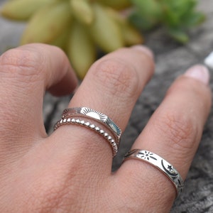 May include: Three silver rings stacked on a finger. The top ring has a sun design, the middle ring is a beaded band, and the bottom ring has a moon and star design.