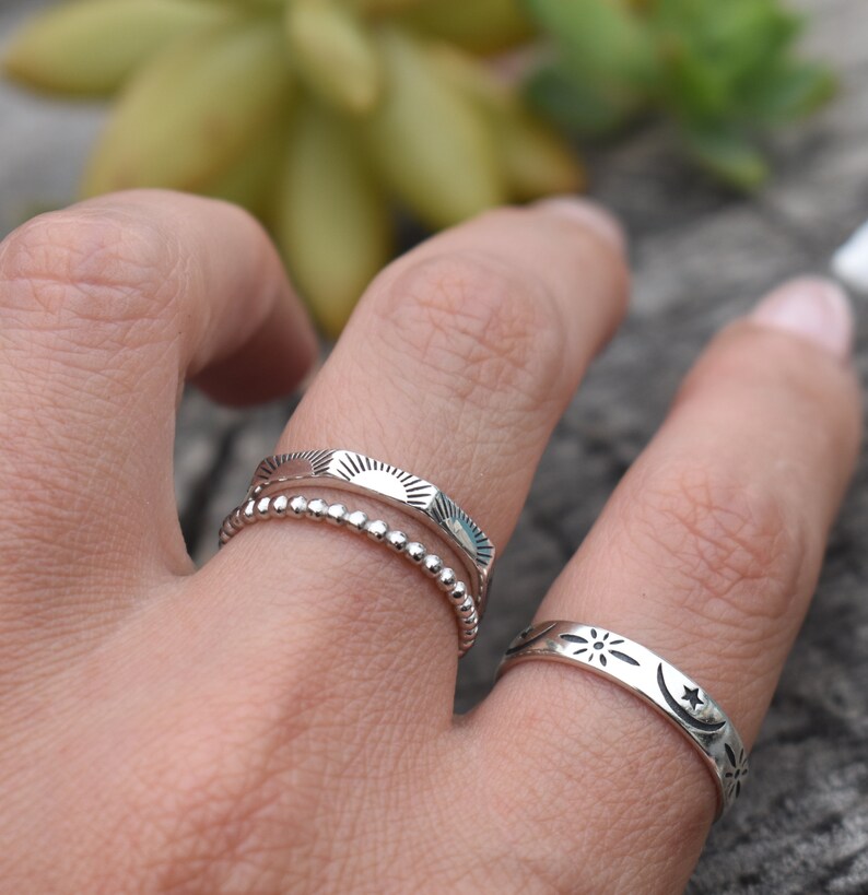 May include: Three silver rings stacked on a finger. The top ring has a sun design, the middle ring is a beaded band, and the bottom ring has a moon and star design.