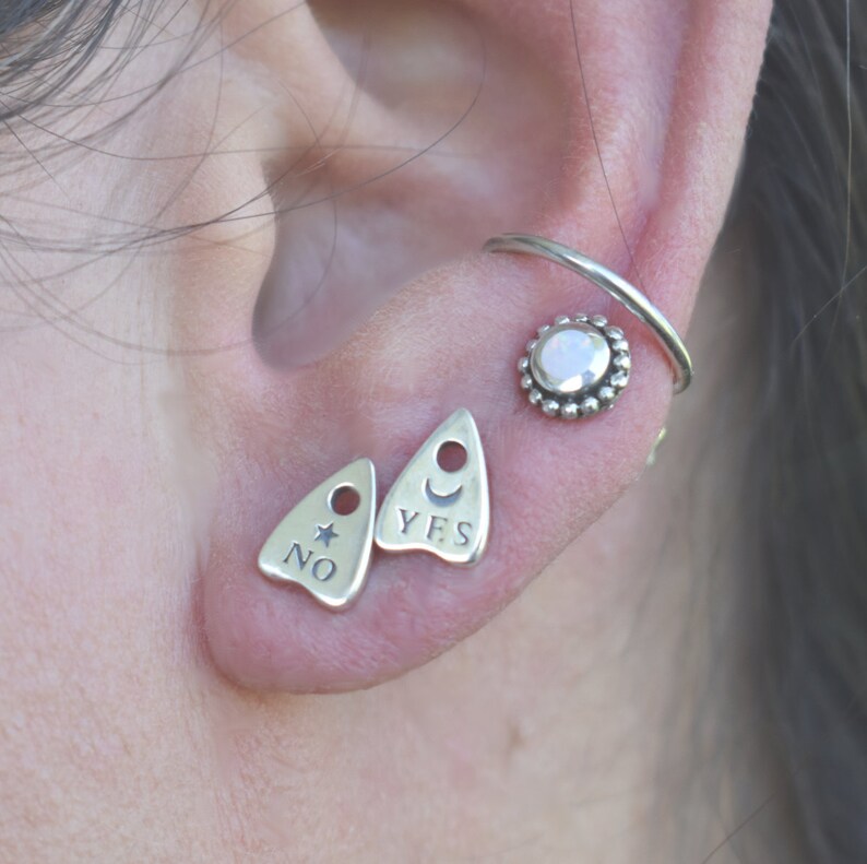 May include: Silver earrings with a crescent moon and star design. The earrings are shaped like a triangle with the words "YES" and "NO" inscribed on them. The earrings are worn in the upper lobe of the ear. A silver hoop earring with a white opal stone is worn in the cartilage of the ear.