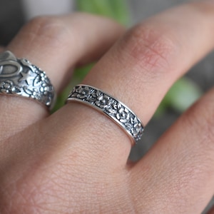May include: Two silver rings with floral designs. The ring on the index finger has a cross and star design. The ring on the middle finger has a floral design.