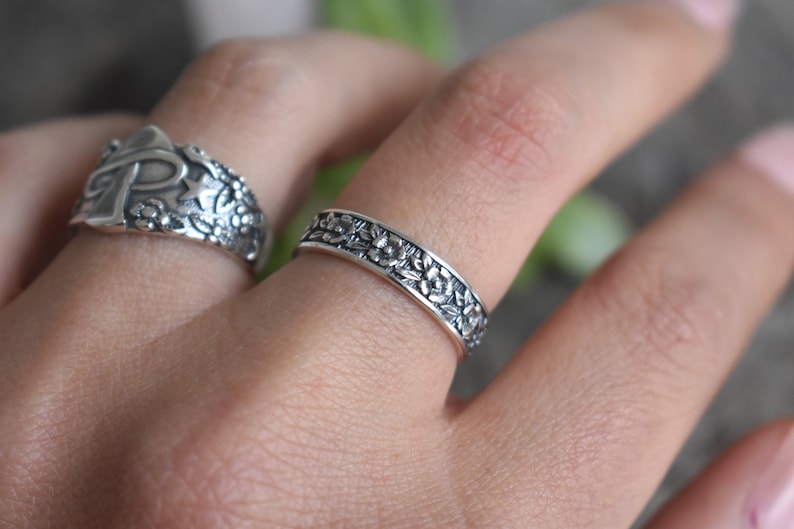 May include: Two silver rings with floral designs. The ring on the index finger has a cross and star design. The ring on the middle finger has a floral design.