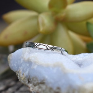 May include: A silver ring with a repeating sun design. The ring is on a light blue rock.