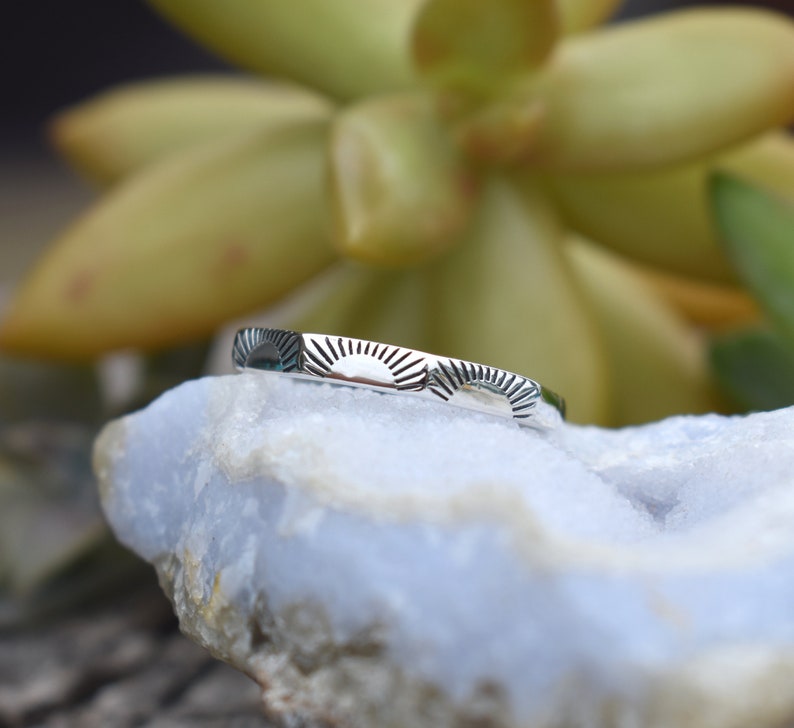 May include: A silver ring with a repeating sun design. The ring is on a light blue rock.