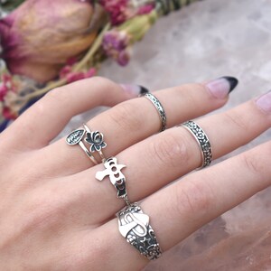 May include: A set of five silver rings with different designs. The rings feature floral patterns, a bird, and a teardrop with a flower. The rings are worn on a hand with a black nail polish.