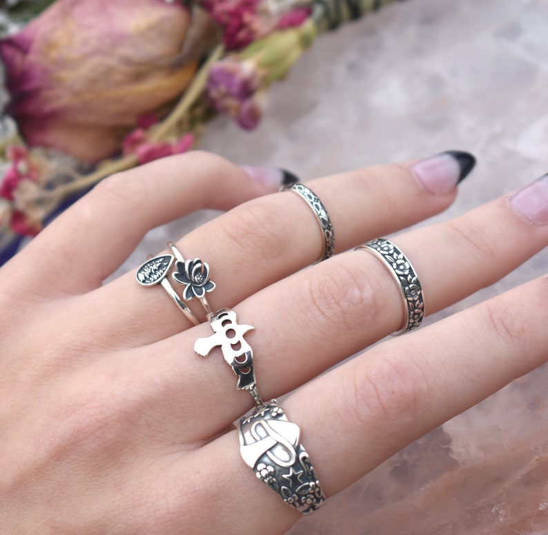 May include: A set of five silver rings with different designs. The rings feature floral patterns, a bird, and a teardrop with a flower. The rings are worn on a hand with a black nail polish.