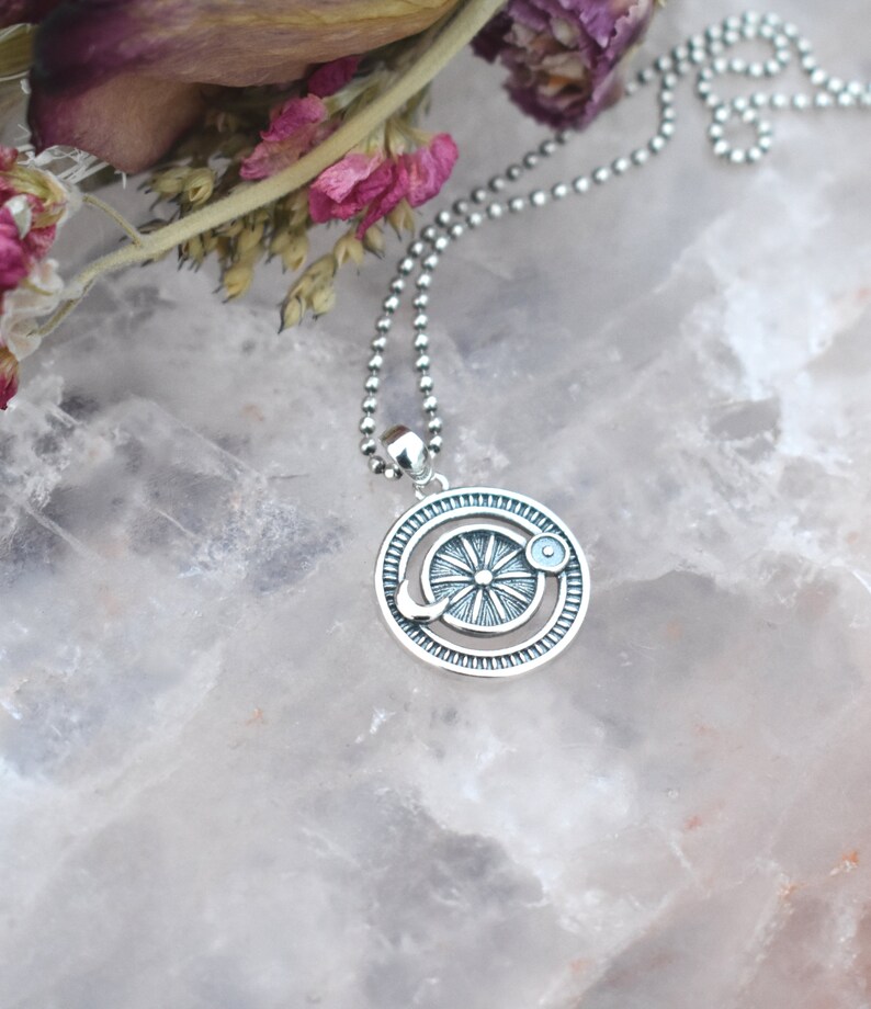 May include: A silver necklace with a circular pendant featuring a sun and moon design. The pendant has a textured, detailed design and is on a silver ball chain.