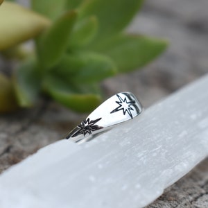 May include: A silver ring with two starburst designs. The ring is on a white surface.
