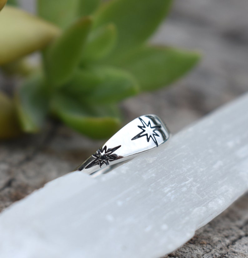 May include: A silver ring with two starburst designs. The ring is on a white surface.