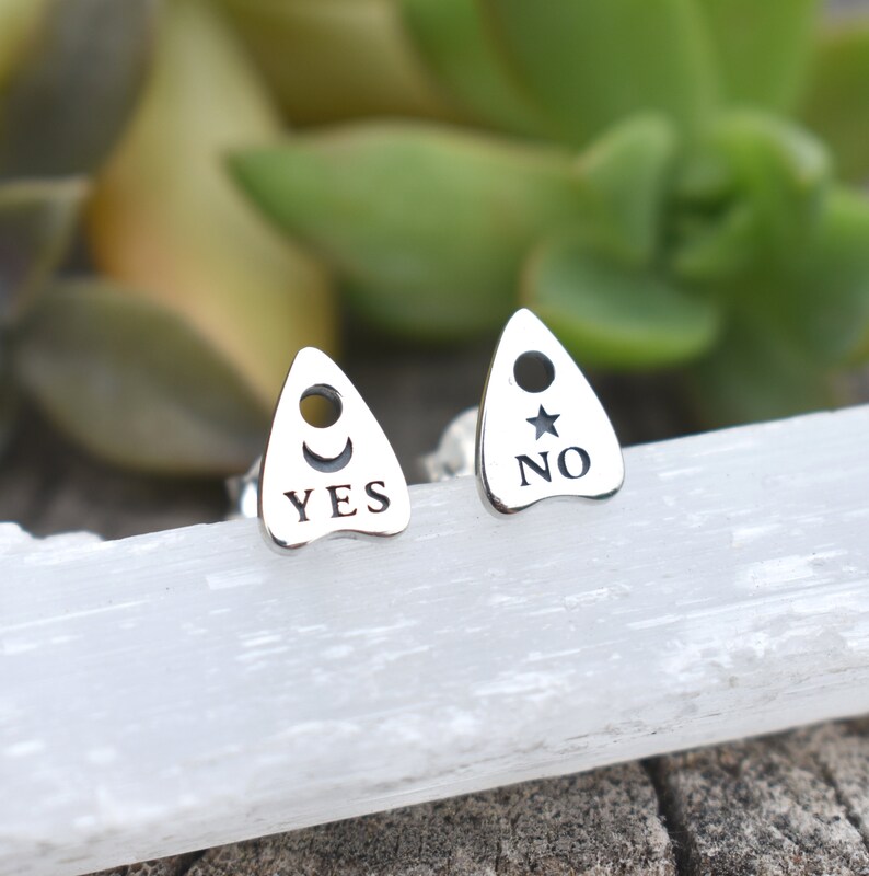 May include: A pair of silver earrings shaped like a Ouija board planchette. One earring says "YES" with a crescent moon and the other says "NO" with a star.