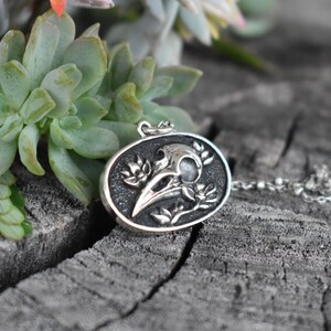 May include: A silver pendant necklace featuring a detailed raven skull with floral accents. The pendant is oval-shaped and hangs from a silver chain.