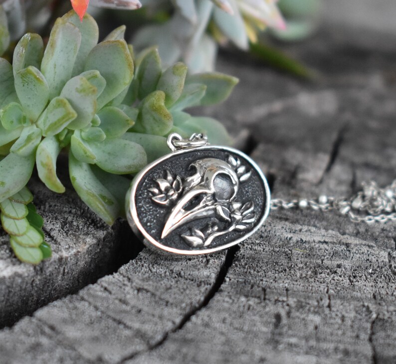 May include: A silver pendant necklace featuring a detailed raven skull with floral accents. The pendant is oval-shaped and hangs from a silver chain.