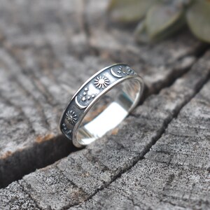 May include: A silver ring with a sun, moon, and stars design. The ring is on a wooden surface.
