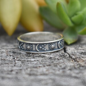 May include: A silver ring with a celestial design featuring moons, stars, and suns. The ring is on a wooden surface.