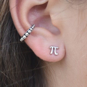 May include: A close-up of a person's ear with two silver earrings. The earring on the lower lobe is a small, silver pi symbol. The earring on the upper lobe is a silver hoop with small, round beads.