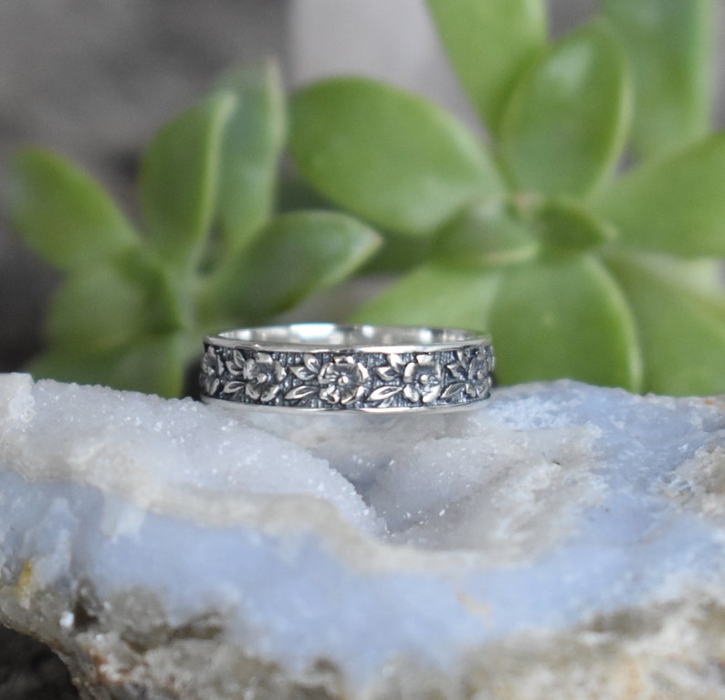 May include: A silver band ring with a floral design. The ring is set against a blue and white background.