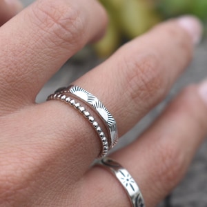 May include: Two silver rings on a finger. The top ring has a textured design of repeating sunbursts. The bottom ring is a simple band of small beads.