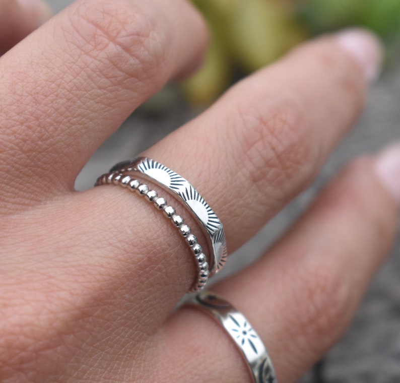 May include: Two silver rings on a finger. The top ring has a textured design of repeating sunbursts. The bottom ring is a simple band of small beads.