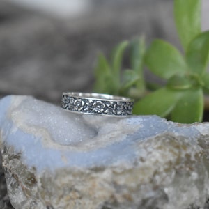 May include: A silver band ring with a floral design. The ring is on a light blue and gray rock.