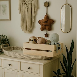 Könnte beinhalten: Eine weiße Kommode mit zwei Schubladen und zwei Türen, eine Wickelunterlage aus Weide, eine Holzkiste mit Stofftieren, ein braunes sternförmiges Kissen mit dem Text "Sweet dreams", ein goldener Spiegel und zwei Topfpflanzen.