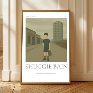 May include: Framed art print featuring the title "SHUGGIE BAIN" with a boy standing on a street. The artwork has a muted color palette with a boy in a dark blue shirt and shorts. The print is in a gold frame.