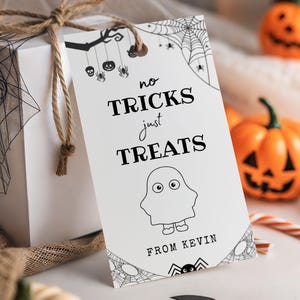 May include: A white gift box tied with twine, featuring a Halloween-themed tag. The tag reads "no tricks just treats" with a ghost illustration and "From Kevin." Decorative pumpkins and a candy cane are in the background.