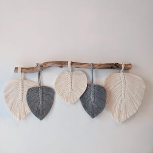 Peut inclure: Une suspension murale avec cinq plumes en macramé, deux grises et trois blanches, suspendues à une branche naturelle.