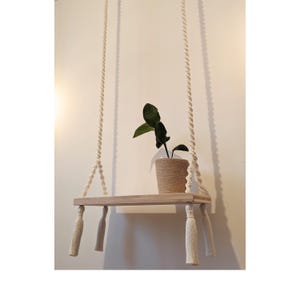 Peut inclure: Étagère suspendue en bois clair, suspendue par des cordes épaisses et blanches. Elle supporte une petite plante en pot aux feuilles vert foncé. Des glands décoratifs ornent les coins, créant une ambiance bohème.