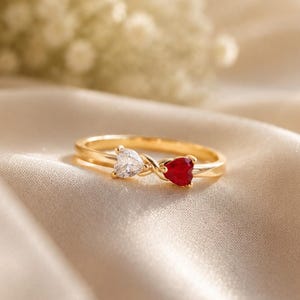 May include: A gold ring featuring two heart-shaped stones, one clear and one red, set in a bypass design. The band is thin and metallic, showcasing a delicate crossover style that holds the two distinct heart-shaped gems securely in place.