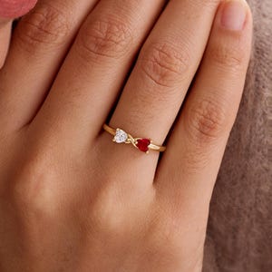 May include: A gold ring featuring two heart-shaped gemstones, one white and one red. The ring's band is thin and yellow, with the hearts set side-by-side. The ring is on a person's finger.