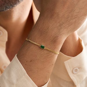 Könnte beinhalten: Ein goldfarbenes Armband mit einem rechteckigen, smaragdgrünen Edelstein. Das Armband hat ein Kettenglied-Design. Das Bild zeigt das Armband am Handgelenk einer Person, mit einem hellen Hemd im Hintergrund.