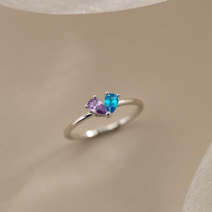 May include: A silver ring featuring two teardrop-shaped gemstones, one purple and one blue. The gemstones are set close together, creating a heart-like design. The ring is displayed on a neutral background.