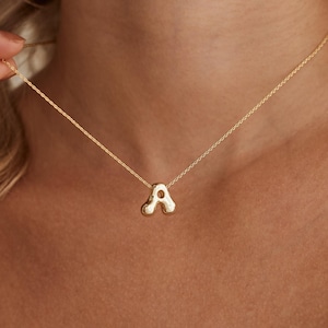 May include: A gold-colored necklace with the letter "A" pendant. The pendant is a stylized, rounded shape. The necklace is on a delicate chain. The image shows the necklace being held by a person.