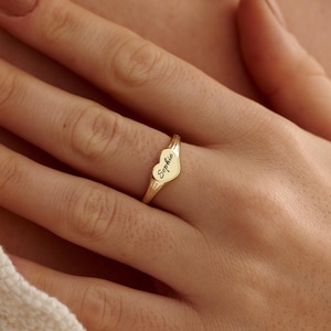 Può includere: Un anello color oro caratterizzato da una parte superiore a forma di cuore con il nome Sophie inciso in corsivo. La fascia è sottile e lucida, con una finitura metallica liscia che avvolge il dito in un design semplice e pulito.