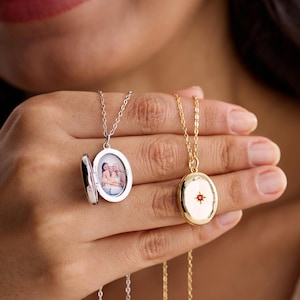Puede incluir: Dos collares con medallones ovalados. Uno es plateado, abierto para revelar una foto. El otro es dorado con un diseño de estrella roja. Ambos están en cadenas finas. La foto muestra a tres personas.