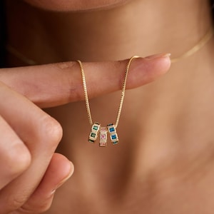 May include: A gold necklace featuring three pendant rings, each set with square gemstones in green, pink, and blue. The necklace is held up by a finger. The chain is a fine, delicate design.