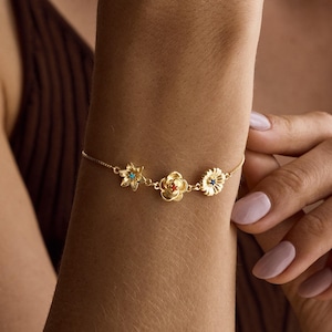 Puede incluir: Una pulsera de cadena en tono dorado con tres dijes florales distintos. El dije de la izquierda es una flor en forma de estrella con un centro turquesa, el del medio es una rosa en capas y el de la derecha es una flor tipo margarita.