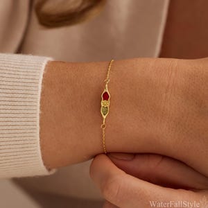 May include: A delicate gold chain bracelet featuring a central pea pod charm. The charm contains three small, circular stones arranged vertically: a deep red stone at the top, a textured yellow stone in the middle, and a mottled green stone at the base.