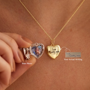 Könnte beinhalten: Eine goldfarbene herzförmige Medaillon-Halskette an einer feinen Kette. Das Medaillon ist geöffnet und zeigt ein kleines Foto einer Person, während die Außenseite mit dem handschriftlichen Text 'Love Daddy xxx' graviert ist. Ein Etikett zeigt auf den Text.