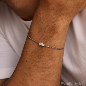 May include: A silver bracelet with a box chain design. The bracelet features a small rectangular charm engraved with the name "ADAM". The bracelet is worn on a person's wrist. The background is a white shirt.
