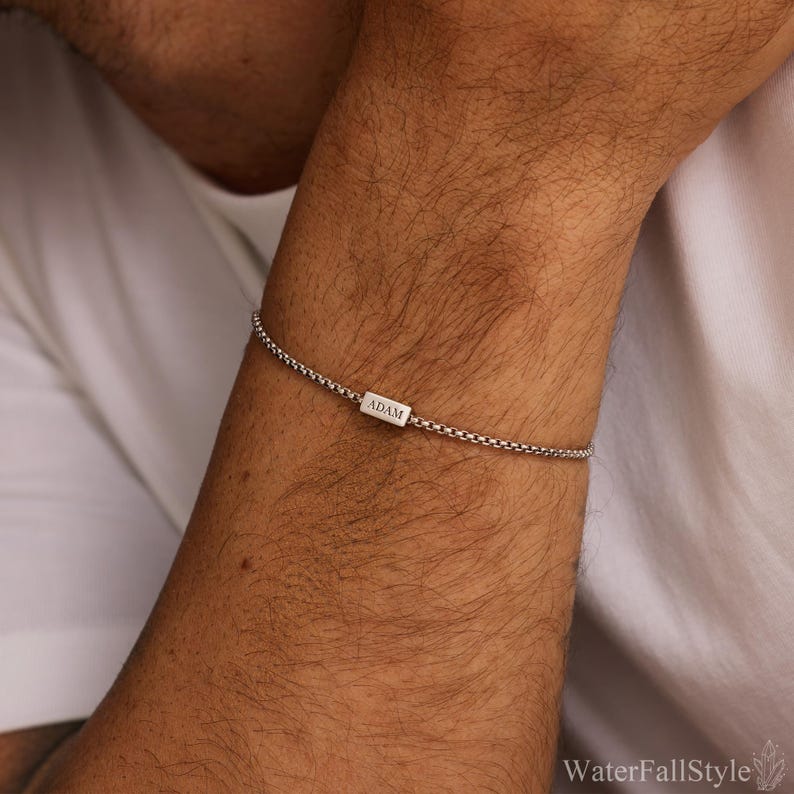 May include: A silver bracelet with a box chain design. The bracelet features a small rectangular charm engraved with the name "ADAM". The bracelet is worn on a person's wrist. The background is a white shirt.