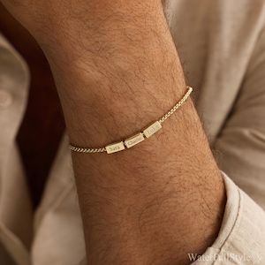 May include: A gold-colored bracelet with three rectangular charms, each engraved with a name: "Sara", "Charles", and "Luke". The bracelet is worn on a person's wrist, and the chain has a delicate, textured design.