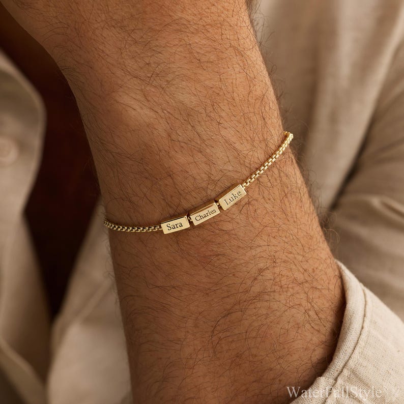 May include: A gold-colored bracelet with three rectangular charms, each engraved with a name: "Sara", "Charles", and "Luke". The bracelet is worn on a person's wrist, and the chain has a delicate, textured design.