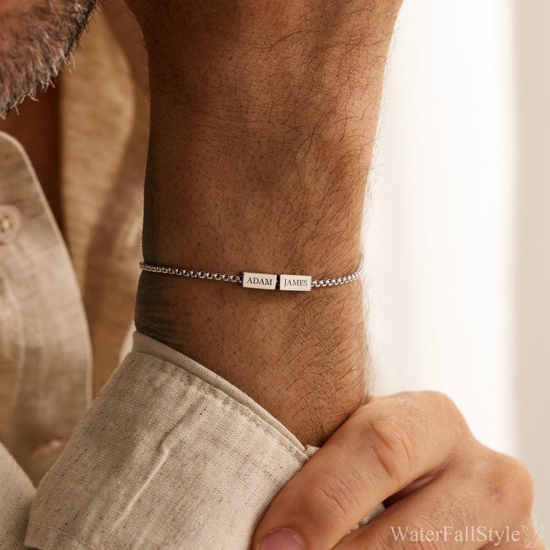 May include: A silver bracelet featuring two rectangular nameplates, each engraved with a name. The bracelet is on a person's wrist, with the names "ADAM" and "JAMES" visible. The bracelet is made of a delicate chain.