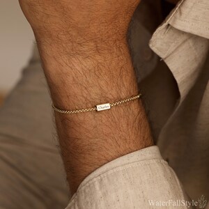 May include: A gold-colored chain bracelet with a rectangular charm. The charm is engraved with the name "Charles". The bracelet is worn on the wrist. The background is a neutral color.