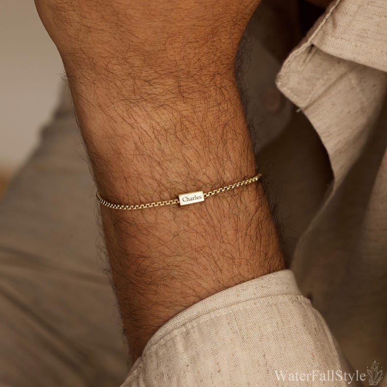 May include: A gold-colored chain bracelet with a rectangular charm. The charm is engraved with the name "Charles". The bracelet is worn on the wrist. The background is a neutral color.