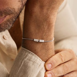 May include: A silver bracelet with a chain link design. The bracelet features a rectangular bar with the engraved names "ADAM" and "JAMES". The bracelet is worn on a wrist.