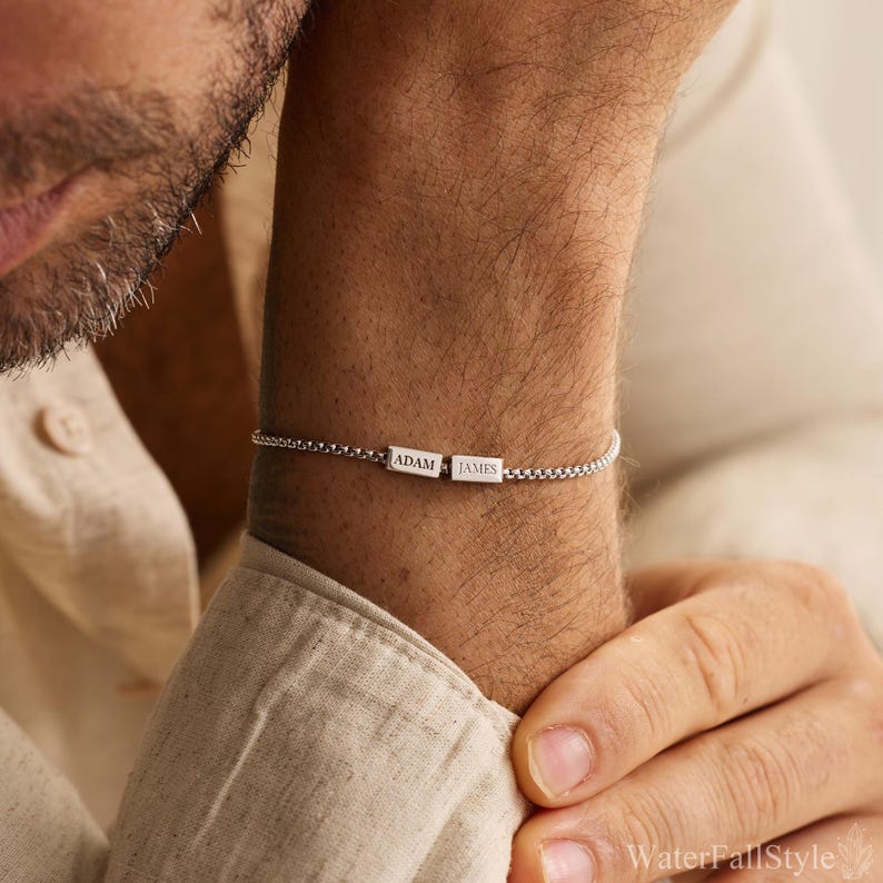 May include: A silver bracelet with a chain link design. The bracelet features a rectangular bar with the engraved names "ADAM" and "JAMES". The bracelet is worn on a wrist.
