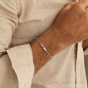May include: A silver bracelet with two rectangular charms, each engraved with a name. The bracelet is a delicate chain style. The background is a neutral beige.