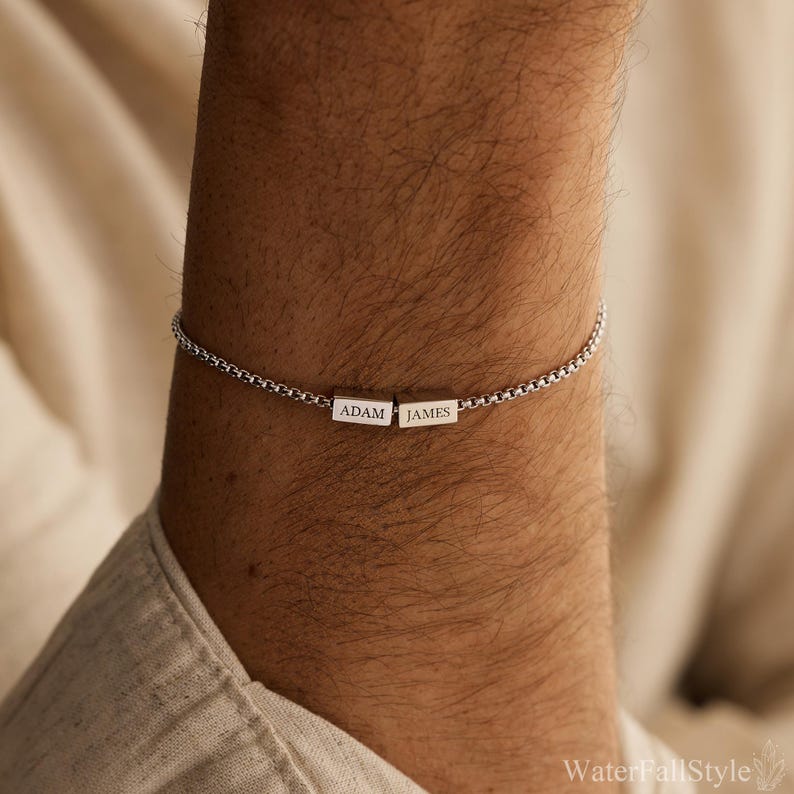 May include: A silver bracelet with a box chain design. Two rectangular silver beads are centered on the bracelet, each engraved with a name: "ADAM" and "JAMES". The bracelet is worn on a person's wrist.